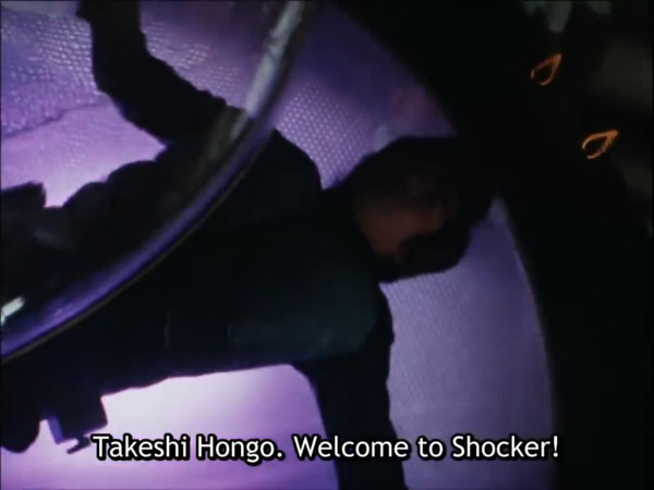 A man in a riding suit is strapped spread-eagle onto a glowing table. A disembodied voice calls out, "Takeshi Hongo. Welcome to Shocker!"